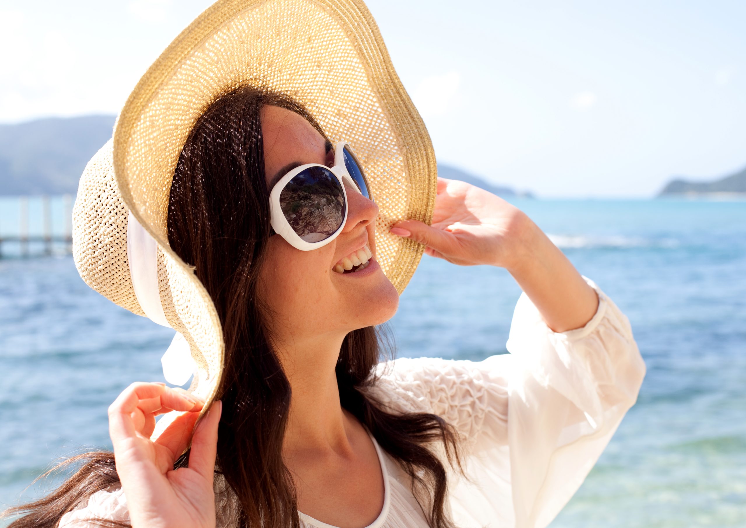 Happy woman wearing summer white dress, hat and sunglasses on beach.